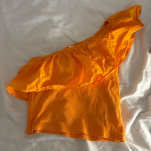 Orange oneshoulder topp med volang - Superfin orange topp med oneshoulder-design och stor volang längs axeln. Toppen är ribbad nedtill och har en snygg croppad passform som verkligen sticker ut. Perfekt för dig som vill ha något färgstarkt och trendigt i garderoben.