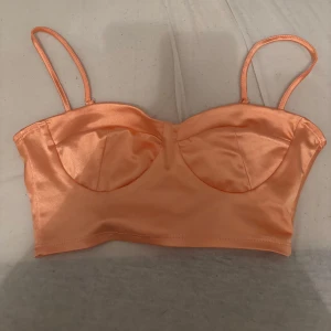 Persikofärgad satin korsett NLY One XS - Snygg persikofärgad korsett-topp från NLY One i satinliknande material. Toppen har tunna axelband och formade kupor för en feminin look. Perfekt att styla med höga jeans eller kjol för en trendig vibe.