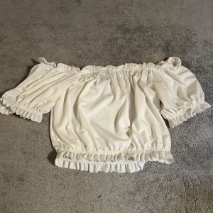 Vit offshoulder topp från Gina Tricot - Säljer en söt vit offshoulder topp från Gina Tricot. Toppen har volangdetaljer vid ärmslut och nederkant, vilket ger en romantisk touch. Perfekt för varmare dagar och kan enkelt matchas med olika outfits.