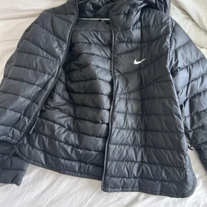 Svart dunjacka från Nike - Säljer en svart dunjacka från Nike med huva och dragkedja. Jackan har en stilren design med det klassiska Nike-logotypen på bröstet. Perfekt för kyligare dagar.