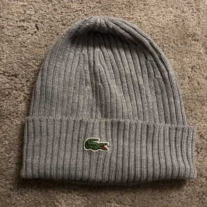 Grå ribbad mössa från Lacoste - Snygg grå ribbad mössa från Lacoste med det klassiska krokodilmärket framtill. Perfekt för kyliga dagar och ger en stilren look. Passar både till vardags och mer avslappnade tillfällen.