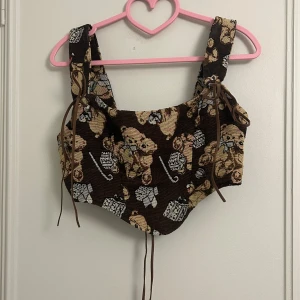 Brun mönstrad korsettopp - Säljer en gullig brun korsettopp med ett charmigt nallemönster. Toppen har breda axelband och snörning på sidorna för justerbar passform. Perfekt för att ge en lekfull touch till din outfit! Skulle säga att den passar s-m<3