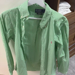 Grön och vitrandig skjorta från Polo Ralph Lauren - Snygg grön- och vitrandig skjorta från Polo Ralph Lauren i slim fit. Skjortan har långa ärmar och knappar framtill. Perfekt för sommaren. Plagget är endast använt en gång!