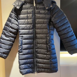 Svart dunjacka från Gant - Säljer en snygg svart dunjacka från Gant med dragkedja och huva. Jackan har en quiltad design och är perfekt för kyligare väder. Den har långa ärmar och en stilren look.