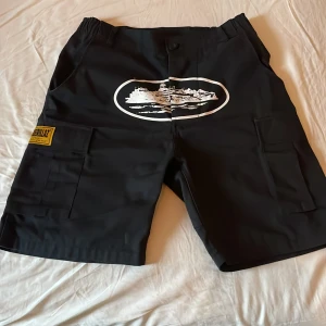 Svarta Cortiez shorts - Snygga svarta shorts med ett coolt tryck på framsidan. De har flera praktiska fickor och en knappstängning. Perfekta för en avslappnad stil. bra för sommaren