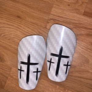 Vita benskydd med korsmotiv - Snygga vita benskydd med ett svart korsmotiv. Perfekta för fotbollsspelare som vill ha stil och skydd på fotbollsplanen.