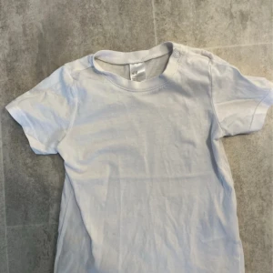 Vit t-shirt från H&M för barn - Säljer en enkel och stilren vit t-shirt från H&M. Den har korta ärmar och en rund halsringning med knappdetalj vid axeln. Perfekt basplagg för alla tillfällen.