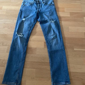 Blå jeans från Jack & Jones - Säljer ett par blå jeans från Jack & Jones med slitna detaljer på benen. De har en klassisk femficksdesign och en knappgylf. Perfekta för en avslappnad stil. Jätte bra skick knappt andvända🙌 kan gå ner i pris vid snabb affär👍
