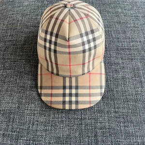 Rutig keps från Burberry - Snygg keps från Burberry med klassiskt rutigt mönster i beige, svart och rött. Kepsen har en justerbar rem baktill för perfekt passform. Perfekt för att ge din outfit en stilren touch. storlek S