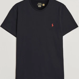 Svart t-shirt från Polo Ralph Lauren - Snygg svart t-shirt från Polo Ralph Lauren med det ikoniska röda broderade logotypen på bröstet. Perfekt för en stilren och avslappnad look. Tillverkad i mjuk bomull för extra komfort.