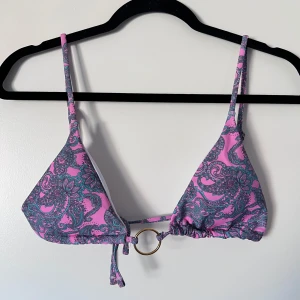 Mönstrad bikiniöverdel med ringdetalj - Snygg bikiniöverdel i rosa , blå och turkos med ett unikt mönster. Den har en dekorativ ringdetalj framtill och justerbara axelband för perfekt passform. Perfekt för sommarens stranddagar! Storlek L men liten i kuporna och passar bra till S/M. Önskar du veta exakta mått? Tveka inte att höra av dig!