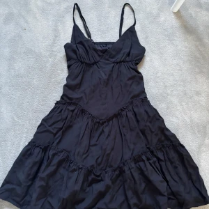 Svart volang klänning med smala axelband - Snygg svart klänning med smala axelband och volangdetaljer. Perfekt för en stilren look. Klänningen har en v-ringad design  i ett mjukt material 