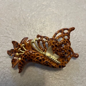 Hårklämma med leopardmönster och fjärilsdetalj - Unik hårklämma i leopardmönster med en elegant guldfärgad fjärilsdetalj. Klämman har en intrikat design med utskurna mönster och är perfekt för att ge en extra touch till din frisyr.