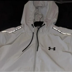 Grå vindjacka från Under Armour - Säljer en grå vindjacka från Under Armour med huva och dragkedja. Jackan har reflekterande detaljer på axlarna och sidfickor. Perfekt för blåsiga dagar.
