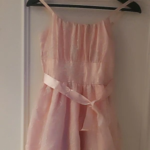 Rosa klänning med spetsdetaljer, storlek 12-13 år - Charmig rosa klänning med spetsdetaljer och ett fint satinband i midjan. Klänningen har smala axelband och en vacker struktur i tyget. Perfekt för speciella tillfällen.