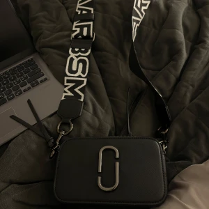 Svart axelväska från Marc Jacobs - Snygg svart axelväska från Marc Jacobs med ett brett axelband med logotyp. Väskan har en stilren design med metallogo på framsidan och dragkedja upptill. Perfekt för att bära dina nödvändigheter med stil. Helt Ny utan defekter eller rost, man kan ha bandet på två sidor om man föredrar svart, skriv för mer bilder eller info, pris kan diskuteras🌸