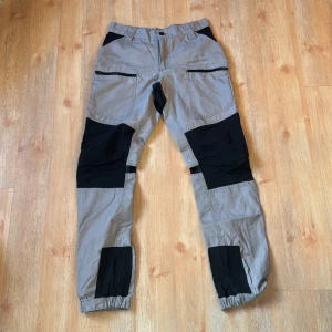 Beige och svarta byxor från Lager 157 - Säljer ett par beige byxor med svarta detaljer från Asci. Byxorna har flera fickor och elastiska muddar vid bensluten. Perfekta för en aktiv livsstil med sin slitstarka design.