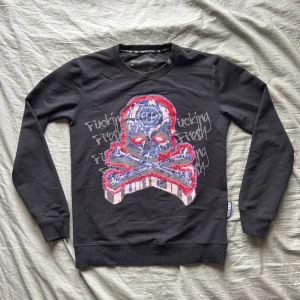 Philipp Plein  - Säljer en svart tröja från Philipp Plein med ett stort dödskallemotiv i rött och blått på framsidan. Texten 'Fucking Fresh' är tryckt både fram och bak. Tröjan har långa armar 