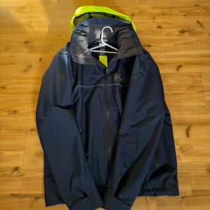 Marinblå regnjacka från Helly Hansen - Säljer en marinblå regnjacka från Helly Hansen med en iögonfallande gul huva. Jackan har dragkedja framtill och långa ärmar. Perfekt för regniga dagar och håller dig torr och bekväm. Är lite stor (inte i längden) på mig som är ca 175-177 och väger 69kg.