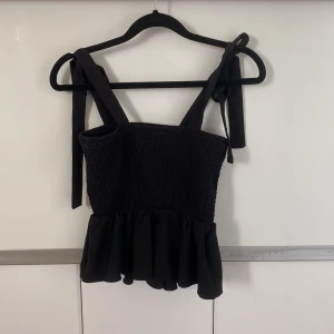 Svart topp från Shein - Snygg svart topp från Shein med smockdetaljer och volang nedtill. Toppen har breda axelband som knyts för en justerbar passform. Perfekt för en stilren look.