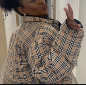 Rutig burrberry pufferjacka - Pufferjacka som är inspirerad av burrberry. En trendig rutig pufferjacka i beige och svart med inslag av rött. Jackan har en oversized passform och långa ärmar. Perfekt för att hålla sig varm och stilig under kyligare dagar. (Defekt: hål i högra fickan)