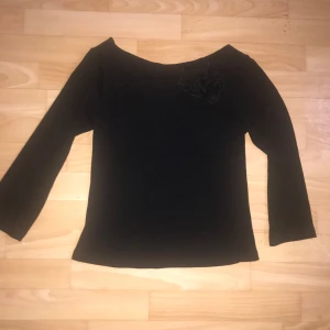 Svart båthals topp med blomdetalj  - Snygg svart topp från CDE med båthalsuringning och en elegant blomdetalj på axeln. Toppen har trekvartsärmar och en stilren design som passar perfekt för olika tillfällen. Väldigt fransk, väldigt flamenco.