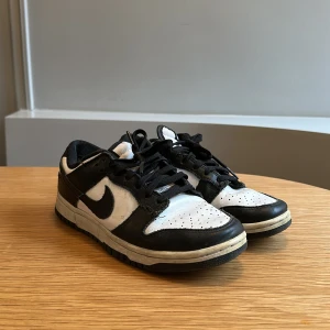 Nike sneakers - Modell: low dunks. Säljer ett par klassiska svartvita Nike sneakers med snörning. Skorna har en stilren design med svart Nike-logga på sidan och perforerad tå för ventilation. Perfekta för en casual look.