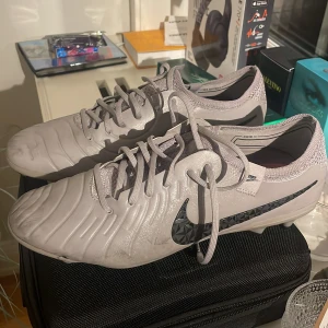 Grå fotbollsskor från Nike - Snygga grå fotbollsskor från Nike med svart logotyp och snörning. Skorna har en modern design, som inte säljs längre på marknaden. Skick 8/10 lite använda men väldigt fina och framförallt hela. Perfekta för fotbollsträning och matcher.