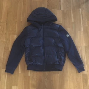 Mörkblå cardigan från Bvaldi - Säljer en mörkblå cardigan från Bvaldi med huva och dragkedja. Jackan har en pufferdesign på framsidan och är tillverkad i syntetmaterial. Perfekt för kyligare dagar med sin stilrena look och praktiska fickor.