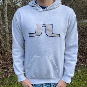 Vit hoodie från J.Lindeberg - Säljer en stilren vit hoodie från J.Lindeberg med ett svart logotyptryck på bröstet. Tröjan har en klassisk känguruficka och ribbade muddar vid ärmslut och nederkant. Perfekt för en avslappnad stil.