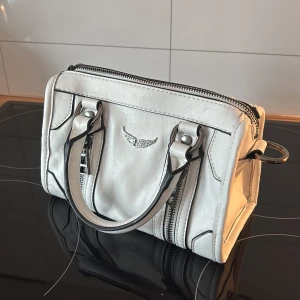 Zadig Vit handväska med dragkedja - Snygg vit handväska med dubbla handtag och coola dragkedjedetaljer. Väskan har en stilren design med ett emblem framtill. Perfekt för att ge din outfit en extra touch.