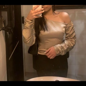 Beige offshoulder topp från NA-KD - Snygg beige offshoulder topp från NA-KD med långa ärmar och en elegant design. Perfekt för en stilren look. Tillverkad i ett glansigt material som ger en lyxig känsla. Används bara en gång.