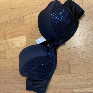 Svart paljettprydd bh från Ella M - Snygg axelbandslös bh från Ella M med glittrande paljetter och en söt rosett framtill. 