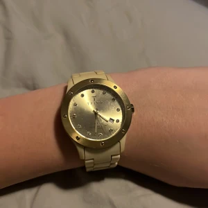 Beige och guld klocka från Triwa har slut på batteri  - Snygg klocka från Triwa med beige armband och guldfärgad urtavla. Urtavlan har små stenar som markörer och ett datumfönster. Perfekt accessoar för att lyfta din stil.