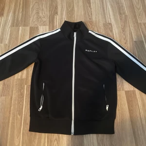 Svart zip från Replay - Snygg svart limited edition zip från Replay med vita ränder längs ärmarna och dragkedja framtill. Jackan har en hög krage och två fickor med dragkedjor. Replay-loggan syns både fram och bak i vitt och grönt. Perfekt för en fräsch och unik look! Pris går att diskutera!