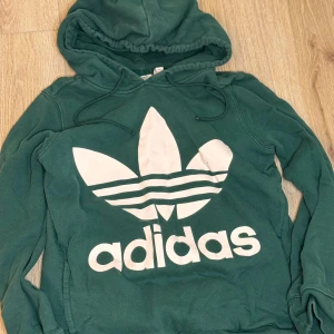 Grön hoodie från Adidas - Säljer en grön hoodie från Adidas med det klassiska logotyptrycket i vitt på framsidan. Tröjan har en bekväm huva med dragsko och långa ärmar. Perfekt för en avslappnad stil.