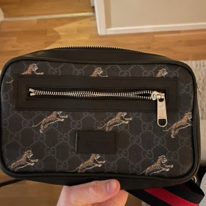 Svart axelväska med tigertryck - Svart axelväska med tigertryck och märkets ikoniska mönster. Väskan har en praktisk dragkedja framtill och en justerbar axelrem i rött och svart. Perfekt för att ge din outfit en lyxig touch.