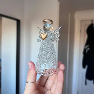 Dekorativ glasängel med belysning - Vacker glasängel med inbyggd belysning som lyser upp i olika färger. Ängeln har ett guldfärgat hjärta och en glittrig design. Perfekt som dekoration för jul eller andra högtider. 🪽🌈 Önskar du veta exakta mått? Tveka inte att höra av dig!