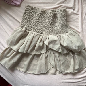 Beige volangkjol från Neo Noir - Säljer en söt beige kjol från Neo Noir med volanger och smockdetaljer i midjan. Kjolen har en luftig och lekfull design som passar perfekt för varmare dagar. Perfekt för dig som vill ha en stilren men ändå trendig look.