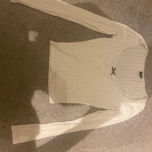 Vit ribbad långärmad topp - Säljer en stilren vit ribbad topp med lång ärm och en söt liten rosett framtill. Perfekt för en enkel och chic look. Toppen har en v-ringad design som ger en feminin touch.