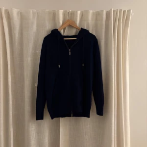 Mörkblå huvtröja med dragkedja - Säljer en stilren mörkblå huvtröja med dragkedja och långa ärmar. Tröjan har en praktisk huva med snörning och en stor ficka framtill. Perfekt för en avslappnad look.