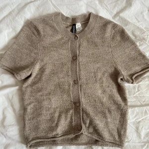 Beige kortärmad kofta från H&M - Säljer en stilren beige kofta från H&M:s Divided-kollektion. Koftan har korta ärmar och knappar framtill, perfekt för lager-på-lager-stil. Den är tillverkad i ett mjukt material som ger en bekväm passform.