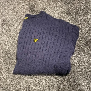 Blå stickad tröja från Lyle & Scott - Säljer en snygg blå stickad tröja från Lyle & Scott med kabelstickat mönster. Tröjan har långa ärmar och ett broderat gult emblem på bröstet. 