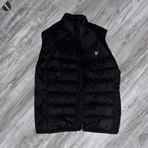 Svart dunväst från Lyle & Scott - Säljer en stilren svart dunväst från Lyle & Scott med dragkedja och ett gult broderat emblem på bröstet. Perfekt för kyligare dagar och enkel att matcha med andra plagg.