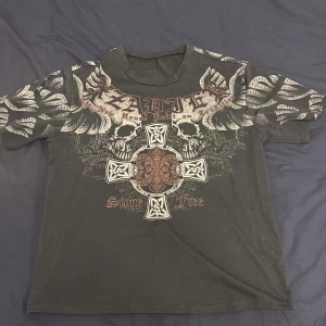 Affliction Tshirt - Säljer en grå t-shirt med ett stort tryck av vingar och dödskallar. Texten 'Stone Free' och ett korsmönster pryder framsidan. Perfekt för en edgy look.