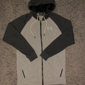Under armour tröja - Säljer en snygg grå och svart hoodie från Under Armour. Den har en dragkedja framtill och en justerbar huva med snören. Perfekt för en sportig look eller avslappnade dagar. Logotypen syns både fram och bak och finns  inga skador.