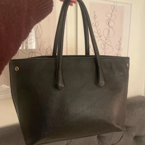 Svart skinnväska - Snygg svart skinnväska med två handtag och stilrena detaljer. Väskan har en rymlig insida med flera fack för enkel organisering. Perfekt för att bära med sig allt man behöver under dagen. En klassisk och tidlös design som passar till många tillfällen.