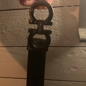Svart bälte med silverdetaljer från Ferragamo - Snyggt svart bälte från Ferragamo med en elegant silverfärgad spänne i form av två sammanlänkade cirklar. Perfekt för att ge en stilren touch till din outfit. Bältet är tillverkat i skinn och har en lyxig känsla.