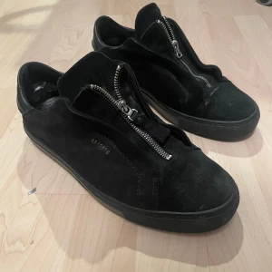 Svarta sneakers med dragkedja från Axel Arigato - Snygga svarta sneakers från Axel Arigato i mocka med dragkedja framtill. Perfekta för en stilren look. Skorna har en låg profil och en diskret logga på sidan. Insidan av skon är skadad eftersom jag har haft inlägg i skorna. Men annars jätte sköna och snygga. Har inte box. Skriv vid frågor eller funderingar!