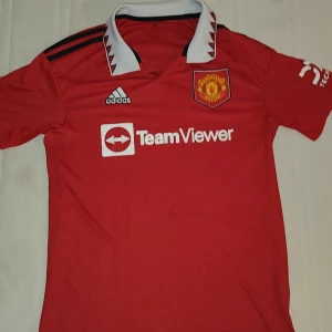 Röd Manchester United fotbollströja från Adidas - Säljer en röd Manchester United fotbollströja från Adidas med vit krage och svarta detaljer. Tröjan har korta ärmar och klubbens emblem på bröstet. Perfekt för fans som vill visa sitt stöd eller bara ha en snygg och stilren fotbollströja att tex träna i eller bara ha den allmänt då den passar till båda och går att matcha med mycket. Den är köpt från Manchester Uniteds officiella hemsida för cirka 800-900
